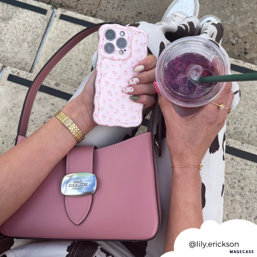 Person holding a Coconut Lane Wavy Phone Case - Ditsy Floral Pink and a purple drink in a clear cup. They have manicured nails and wear a gold watch, bracelet, and rings. A pink Coach handbag rests on their lap. The background is blurred, focusing on accessories. Instagram handle @lily.erickson.