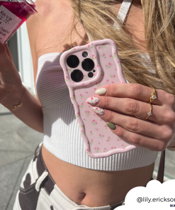A person with long blonde hair is holding a phone adorned with Coconut Lane's Wavy Phone Case - Ditsy Floral Pink in one hand and a jar of a pink beverage in the other. They are wearing a white crop top and have pastel, floral-patterned nails. Their gold jewelry includes a ring and bracelet. The username 