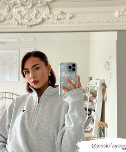 A woman stands in front of a mirror taking a selfie with her phone, adorned in the Pearls & Bows Phone Case by Coconut Lane. She is wearing a light grey zip-up sweatshirt and has long dark hair. Behind her, there is a decorated bedroom with framed pictures on the wall and a vanity with a mirror. Her Instagram handle, @jessiefayeeee, is written on the image.
