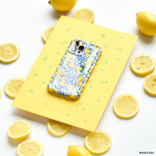 A Coconut Lane Wavy Phone Case - Amalfi Lemon sits on a yellow sheet of paper decorated with green leaf patterns. Surrounding the paper are whole and sliced lemons, creating a bright and cheerful scene.