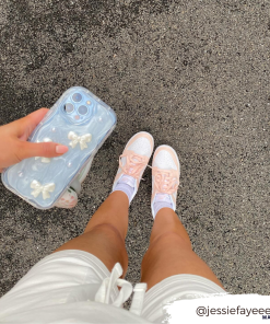 A person's legs are shown from above, wearing white shorts, white socks, and pastel pink sneakers. They are holding a phone with the Pearls & Bows Phone Case by Coconut Lane. The ground is an asphalt surface. The Instagram handle @jessiefayeeee is overlaid in the bottom right corner.