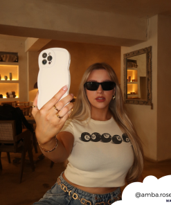 A blonde woman with long hair is taking a selfie with an iPhone encased in a lightweight, highly protective NAKD Curvy Phone Case - Vanilla by Coconut Lane. She is wearing black sunglasses, a white crop top with black text, and blue jeans while standing in a stylish, warmly-lit room. The Instagram handle 