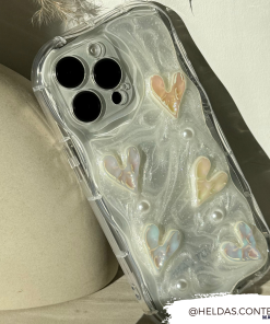 A close-up shot of an iPhone15 enclosed in Coconut Lane's Pearly Hearts Phone Case - Clear, which features sparkling pastel, heart-shaped decorations. The sturdy Pearly Hearts Phone Case lies on a light-colored, textured background. The Instagram handle @HELDAS.CONTENT is displayed in the bottom right corner.