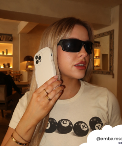 A woman with long blonde hair, wearing dark sunglasses and a white shirt with black graphic letters, holds a huge prop of an iPhone to her ear as if she's talking on it. Behind her is a softly lit room with shelves and a framed mirror. Swipe to shop @amba.rose's favorite NAKD Curvy Phone Case - Vanilla by Coconut Lane—stylish and highly protective!