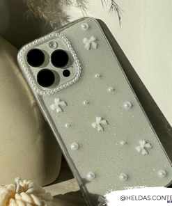 A close-up of a glittery, clear design Mini White Bows Phone Case for iPhone 15 by Coconut Lane, adorned with small white bows and pearls. The phone is leaning against a neutral-colored vase. The Instagram handle 