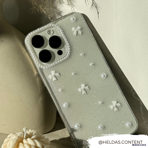 A close-up of a glittery, clear design Mini White Bows Phone Case for iPhone 15 by Coconut Lane, adorned with small white bows and pearls. The phone is leaning against a neutral-colored vase. The Instagram handle "@HELDAS.CONTENT" is displayed in the bottom-right corner of the image.