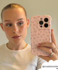 A person with their blonde hair tied back takes a mirror selfie. They are wearing a white top and holding a phone with the Wavy Phone Case - Ditsy Floral Pink by Coconut Lane. The Instagram handle @maddiewildeway is visible in the bottom right corner.