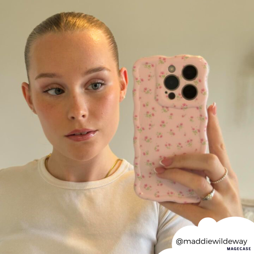 A person with their blonde hair tied back takes a mirror selfie. They are wearing a white top and holding a phone with the Wavy Phone Case - Ditsy Floral Pink by Coconut Lane. The Instagram handle @maddiewildeway is visible in the bottom right corner.