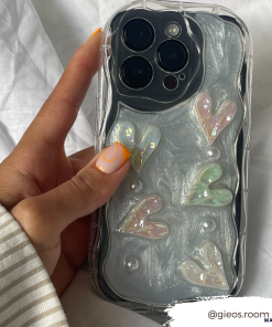 A hand showcasing stylish, swirled orange and white nail art holds an iPhone 15 with the Pearly Hearts Phone Case - Clear from Coconut Lane. This extra protective case features iridescent pearly hearts and a few pearls on a glossy silver background. A white tag at the bottom right corner reads 