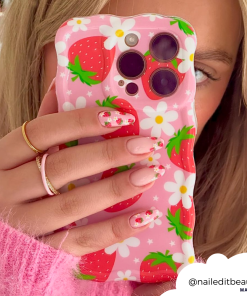 Close-up of a person holding a phone with Coconut Lane's Wavy Phone Case - Berry Cute Strawberry, featuring an adorable strawberry and daisy-themed design. The phone partially covers their face, perfectly matching their strawberry and floral nail art. They are wearing a pink sweater and gold rings. "@naileditbeauty" is displayed in a cloud shape.