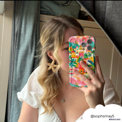 A person with wavy blond hair wearing a white top and starfish-shaped earrings is taking a mirror selfie with their smartphone. The phone case, an eye-catching and colorful design from Coconut Lane called the Wavy Phone Case - Portofino Dreams, features a beautiful Italian seaside motif. The Instagram handle "@sophsmay5" is visible in the bottom right corner.