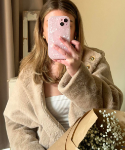A person is taking a mirror selfie with an iPhone 15 that has a Coconut Lane Moonstone Pink Phone Case adorned with flowers. They are wearing a beige faux fur coat over a white top and holding a brown paper-wrapped bouquet of baby's breath flowers. The background shows a curtain and a piece of furniture.