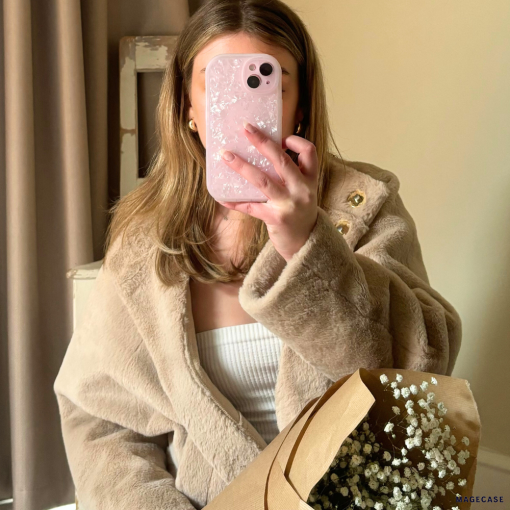 A person is taking a mirror selfie with an iPhone 15 that has a Coconut Lane Moonstone Pink Phone Case adorned with flowers. They are wearing a beige faux fur coat over a white top and holding a brown paper-wrapped bouquet of baby's breath flowers. The background shows a curtain and a piece of furniture.