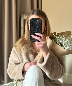 A person with long, light brown hair is sitting indoors and wearing a beige coat. They are holding a bouquet of white flowers wrapped in brown paper in one hand and covering their face with a Moonstone Black Phone Case from Coconut Lane in the other hand. A beige curtain and a wall are in the background.