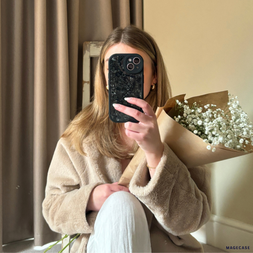A person with long, light brown hair is sitting indoors and wearing a beige coat. They are holding a bouquet of white flowers wrapped in brown paper in one hand and covering their face with a Moonstone Black Phone Case from Coconut Lane in the other hand. A beige curtain and a wall are in the background.