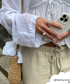 A person wearing a white long-sleeve blouse and beige drawstring pants holds an iPhone adorned with the Wavy Phone Case - Ditsy Floral White from Coconut Lane in both hands. They have painted nails and wear rings. A woven straw bag is slung over their shoulder, and their Instagram handle, @libertycarlyle_, is at the bottom right corner.