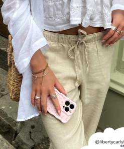 A person wearing beige linen pants and a white long-sleeve top knotted at the waist stands on a stone-paved street. They hold a phone encased in Coconut Lane's Wavy Phone Case - Ditsy Floral Pink, featuring multiple cameras, and have various rings, bracelets, and a straw bag slung over their shoulder. (Credit: @libertycarlyle_).