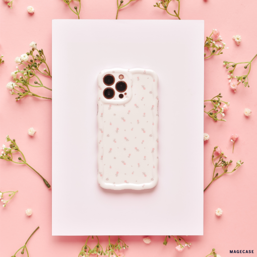 A smartphone encased in Coconut Lane's Wavy Phone Case - Ditsy Floral White lies on a white rectangular sheet, surrounded by delicate sprigs of small white flowers on a pink background. The overall aesthetic is soft and feminine.