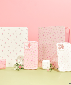 A display of floral-themed tech accessories includes two tablet cases, four iPhone cases (featuring the Wavy Phone Case - Ditsy Floral White by Coconut Lane), and two AirPods cases. The items are arranged on a light green surface against a pink backdrop, with small white flowers accentuating the display.
