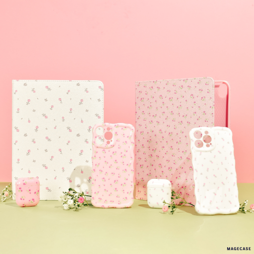 A display of floral-themed tech accessories includes two tablet cases, four iPhone cases (featuring the Wavy Phone Case - Ditsy Floral White by Coconut Lane), and two AirPods cases. The items are arranged on a light green surface against a pink backdrop, with small white flowers accentuating the display.
