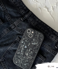 A sleek black smartphone with a textured, crinkled-looking back cover rests in the front pocket of black jeans. The jeans and phone are set against a white textured background. For durable protection, consider the Moonstone Black Phone Case from Coconut Lane. The Instagram handle @_i.xa is visible in the bottom right corner.