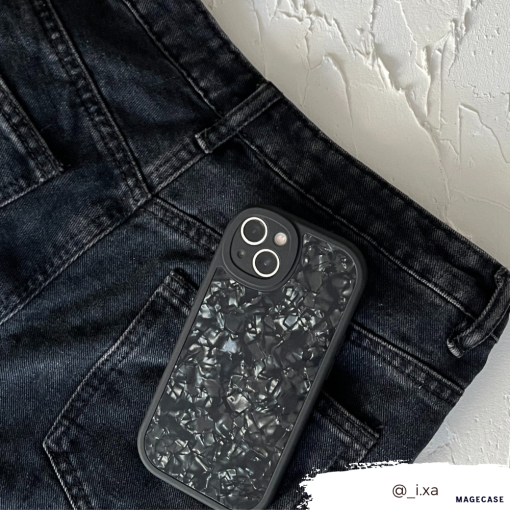 A sleek black smartphone with a textured, crinkled-looking back cover rests in the front pocket of black jeans. The jeans and phone are set against a white textured background. For durable protection, consider the Moonstone Black Phone Case from Coconut Lane. The Instagram handle @_i.xa is visible in the bottom right corner.
