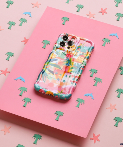 The Wavy Phone Case - Portofino Dreams by Coconut Lane, with its Italian seaside theme and colorful pastel patterns, is placed on a pink surface adorned with small illustrations of palm trees, starfish, and dolphins. This extra protective case adds a lively touch to the overall beachy design.
