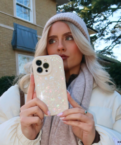 A person with long blonde hair, wearing a beige beanie, scarf, and white jacket, holds a Moonstone Pearl Phone Case by Coconut Lane in front of their face. They are standing outdoors in front of a house with green shrubs and trees in the background.