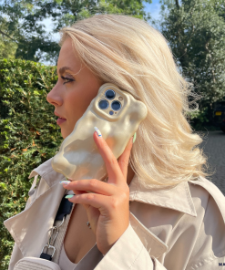 A person with platinum blonde hair holds an iPhone15 with a 3D Pearl Phone Case - Gold by Coconut Lane to their ear. They are wearing a beige trench coat and carrying a small black purse. The background features green foliage and a pathway in a suburban area.