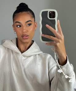 A person with their hair in a bun takes a mirror selfie. They are wearing a light-colored hooded sweatshirt and holding an iPhone 15 with Coconut Lane's Curvy Phone Case in Cream, which is highly protective and has a decorative charm. The background is plain and neutral.