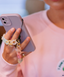 A person wearing a pink sweatshirt from the NAKD spring collection holds their lilac NAKD Phone Case by Coconut Lane, adorned with a colorful chain strap. The phone's full camera protection and finger grip are visible, while the individual's face is partially out-of-frame, concentrating on their pastel favorite.