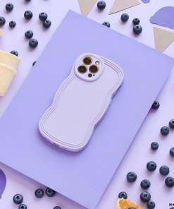 A smartphone in a lightweight, blue NAKD Curvy Phone Case by Coconut Lane rests atop a light purple sheet on a flat surface. Scattered around are blueberries, pieces of waffle cones, and cut-out shapes of ice cream cones and scoops in purple, brown, and white. The scene has a playful and colorful aesthetic.