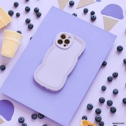 A smartphone in a lightweight, blue NAKD Curvy Phone Case by Coconut Lane rests atop a light purple sheet on a flat surface. Scattered around are blueberries, pieces of waffle cones, and cut-out shapes of ice cream cones and scoops in purple, brown, and white. The scene has a playful and colorful aesthetic.