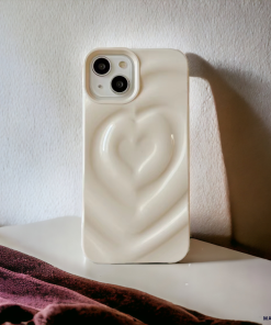A white smartphone with a Melting Heart Phone Case - Coconut Cream by Coconut Lane stands upright on a white surface. A reddish-purple fabric is visible in the foreground, casting shadows. Light enters from the right, illuminating the phone and casting a shadow on the wall behind.