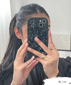 A person with dark hair in a ponytail takes a mirror selfie. They are holding an iPhone15 with the Moonstone Black Phone Case from Coconut Lane, which offers durable protection and partially covers their face. They wear a black top and several rings on their fingers. The Instagram handle @_i.xa is visible in the bottom right corner.