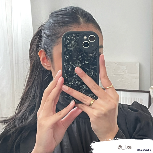 A person with dark hair in a ponytail takes a mirror selfie. They are holding an iPhone15 with the Moonstone Black Phone Case from Coconut Lane, which offers durable protection and partially covers their face. They wear a black top and several rings on their fingers. The Instagram handle @_i.xa is visible in the bottom right corner.