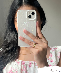 A woman with long black hair holds a white phone with the Coconut Lane Mother of Pearl Phone Case in front of her face, obscuring it. She is wearing a white dress adorned with pink floral patterns and multiple gold rings. The background is neutral, and the handle @_i.xa appears in the bottom right corner.