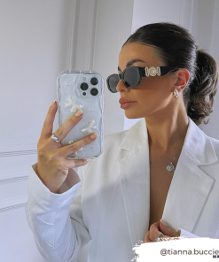A woman with a sleek ponytail takes a mirror selfie while wearing black sunglasses, a white blazer, and minimal jewelry. She holds an iPhone15 with the Coconut Lane Pearls & Bows Phone Case, decorated with white floral designs. The Instagram handle 
