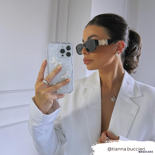 A woman with a sleek ponytail takes a mirror selfie while wearing black sunglasses, a white blazer, and minimal jewelry. She holds an iPhone15 with the Coconut Lane Pearls & Bows Phone Case, decorated with white floral designs. The Instagram handle "@tianna.buccieri" is displayed in the bottom right corner.