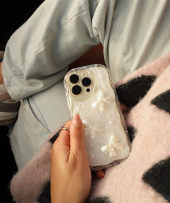 An individual is holding an iPhone 15 adorned with the Pearls & Bows Phone Case by Coconut Lane. They are dressed in light-wash jeans, with a light-colored, fuzzy blanket featuring black spots draped over their legs. Sneakers can be seen near the bottom left—a perfect blend of style and comfort.