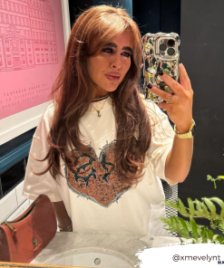 A person with long brown hair takes a mirror selfie in a stylish indoor setting. They wear a white shirt with a detailed design and hold their phone, which is adorned with Coconut Lane's 3D Chrome Phone Case. A pink handbag and some greenery are visible in the foreground. Text in the image reads 