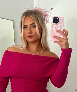 A blonde woman is taking a mirror selfie with her Coconut Lane Melting Heart Phone Case in Peony Pink. She is dressed in an off-the-shoulder, long-sleeve pink top and accessorized with a pink feather in her hair. The background features a plain, white wall and a mirror.