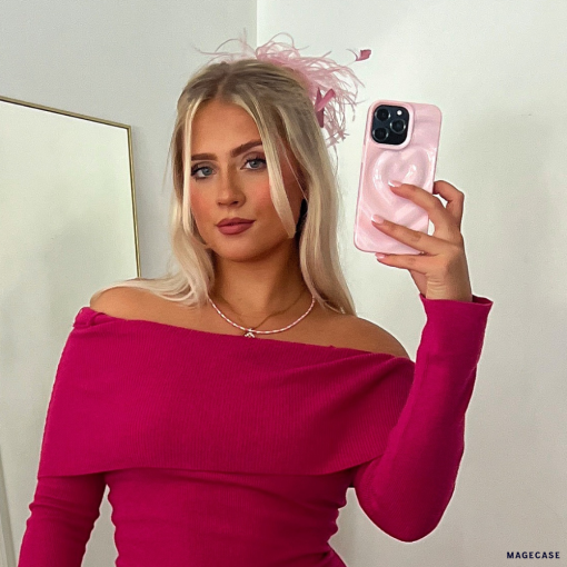 A blonde woman is taking a mirror selfie with her Coconut Lane Melting Heart Phone Case in Peony Pink. She is dressed in an off-the-shoulder, long-sleeve pink top and accessorized with a pink feather in her hair. The background features a plain, white wall and a mirror.