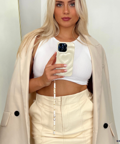 A woman with long blonde hair holds up her phone, taking a mirror selfie. She is wearing a white cropped tank top, a light beige skirt, and a matching beige coat draped over her shoulders. Her phone sports the stylish Melting Heart Phone Case - Coconut Cream by Coconut Lane. The background is plain white, with a dry plant partially visible on the left.