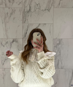 A person stands in front of a marble wall, taking a mirror selfie with a phone adorned in the highly protective NAKD Curvy Phone Case - Vanilla by Coconut Lane. They are wearing a cream-colored cable knit sweater with a white ruffled collar and lace details. Only their torso and part of their head are visible, with the phone covering their face.