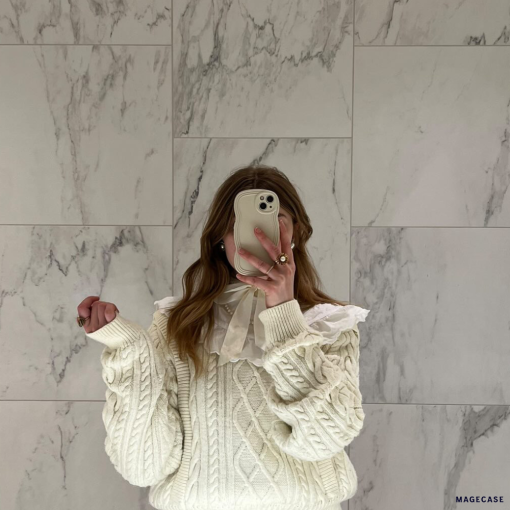 A person stands in front of a marble wall, taking a mirror selfie with a phone adorned in the highly protective NAKD Curvy Phone Case - Vanilla by Coconut Lane. They are wearing a cream-colored cable knit sweater with a white ruffled collar and lace details. Only their torso and part of their head are visible, with the phone covering their face.