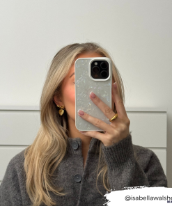 A person with long, blonde hair is taking a mirror selfie with their smartphone, which is adorned with the light-colored Mother of Pearl Phone Case by Coconut Lane. They are wearing a grey sweater and gold hoop earrings. The Instagram handle 