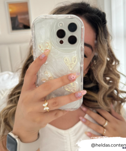 A woman with wavy blonde hair holds up her iPhone15 encased in the Coconut Lane Pearly Hearts Phone Case - Clear, featuring elegant heart designs, taking a selfie. She has floral nail art and wears a ring with a butterfly design. The background includes a white bed and two framed pictures. Text: 
