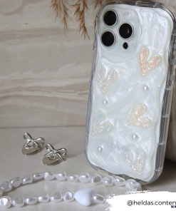 Displayed against a marble backdrop is the Pearly Hearts Phone Case - Clear by Coconut Lane, embellishing an iPhone15 with its pearlescent white casing adorned with shiny heart shapes and pearls. Silver butterfly earrings and a beaded white bracelet accompany the phone. In the bottom right corner, 