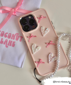 The Pearly Hearts Phone Case - Pink from Coconut Lane is a protective accessory for your iPhone 15, adorned with decorative pearl hearts and pink ribbons. It is displayed next to a pink pouch labeled 